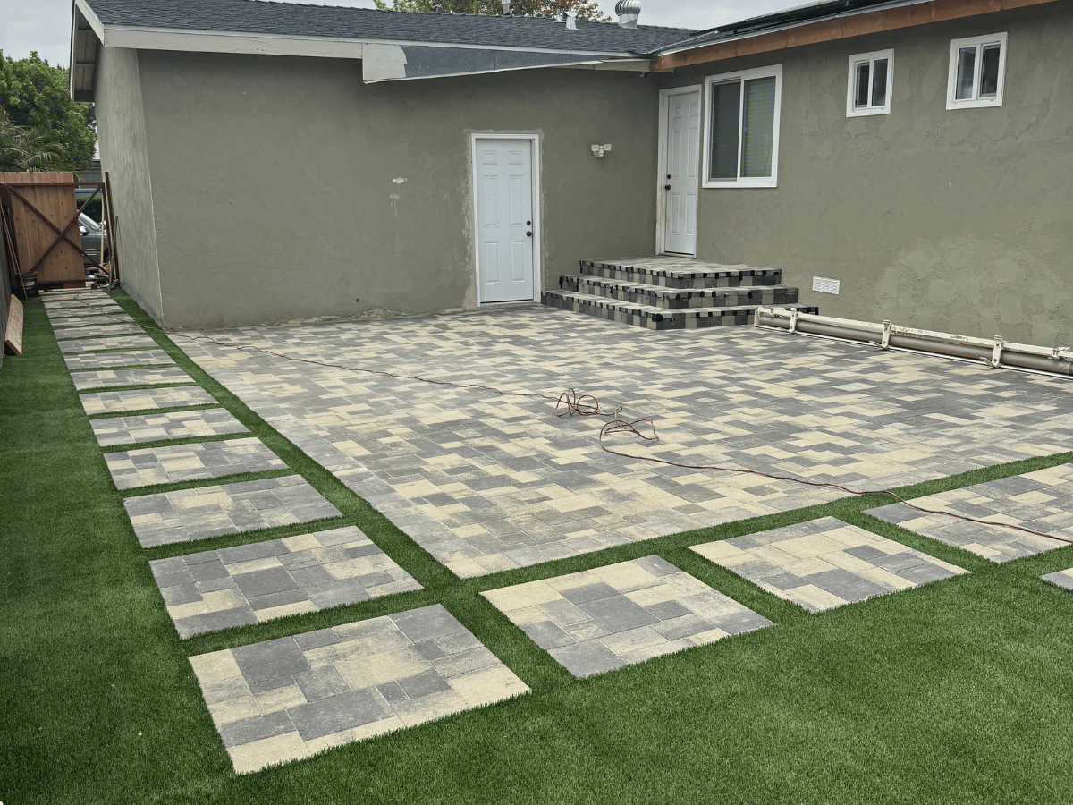 Backyard paver patio in gray and tan blend with artificial turf inlays and paver entry steps