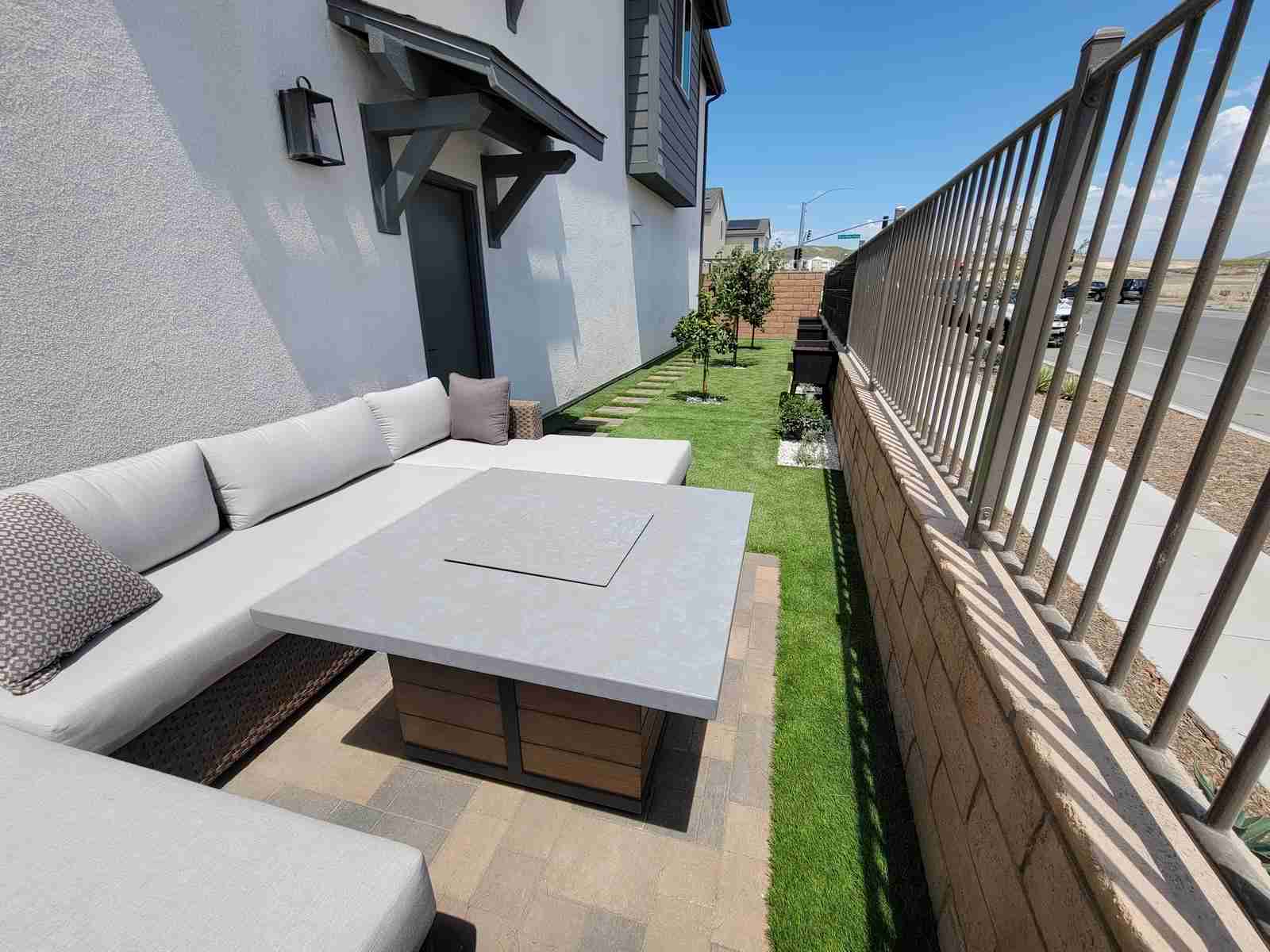 Paver patio with outdoor sectional and fire pit table beside turf, block retaining wall and metal fence