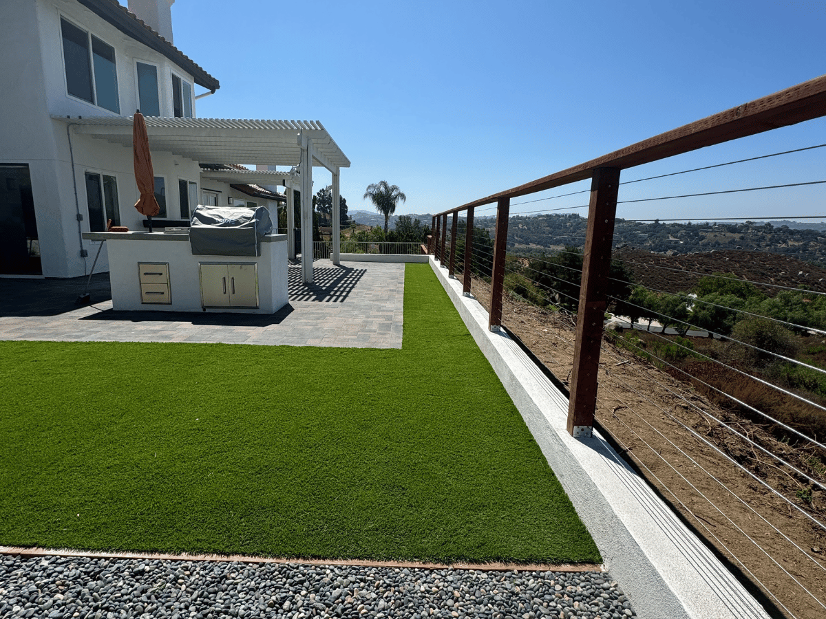 Hillside backyard with artificial turf lawn, paver patio, outdoor kitchen island, and wood-post cable railing