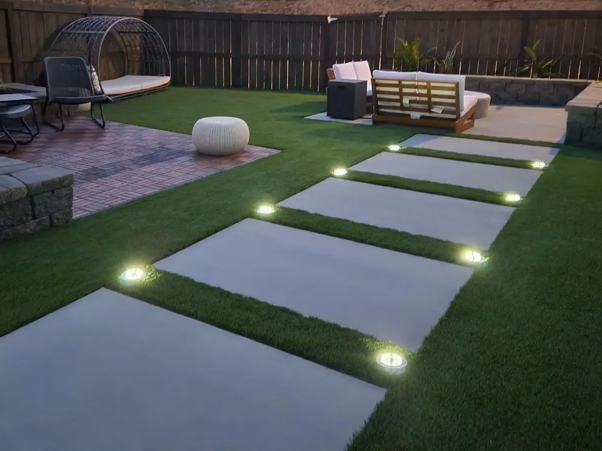 Hillside backyard at dusk with turf, lighted concrete pads and paver seating areas