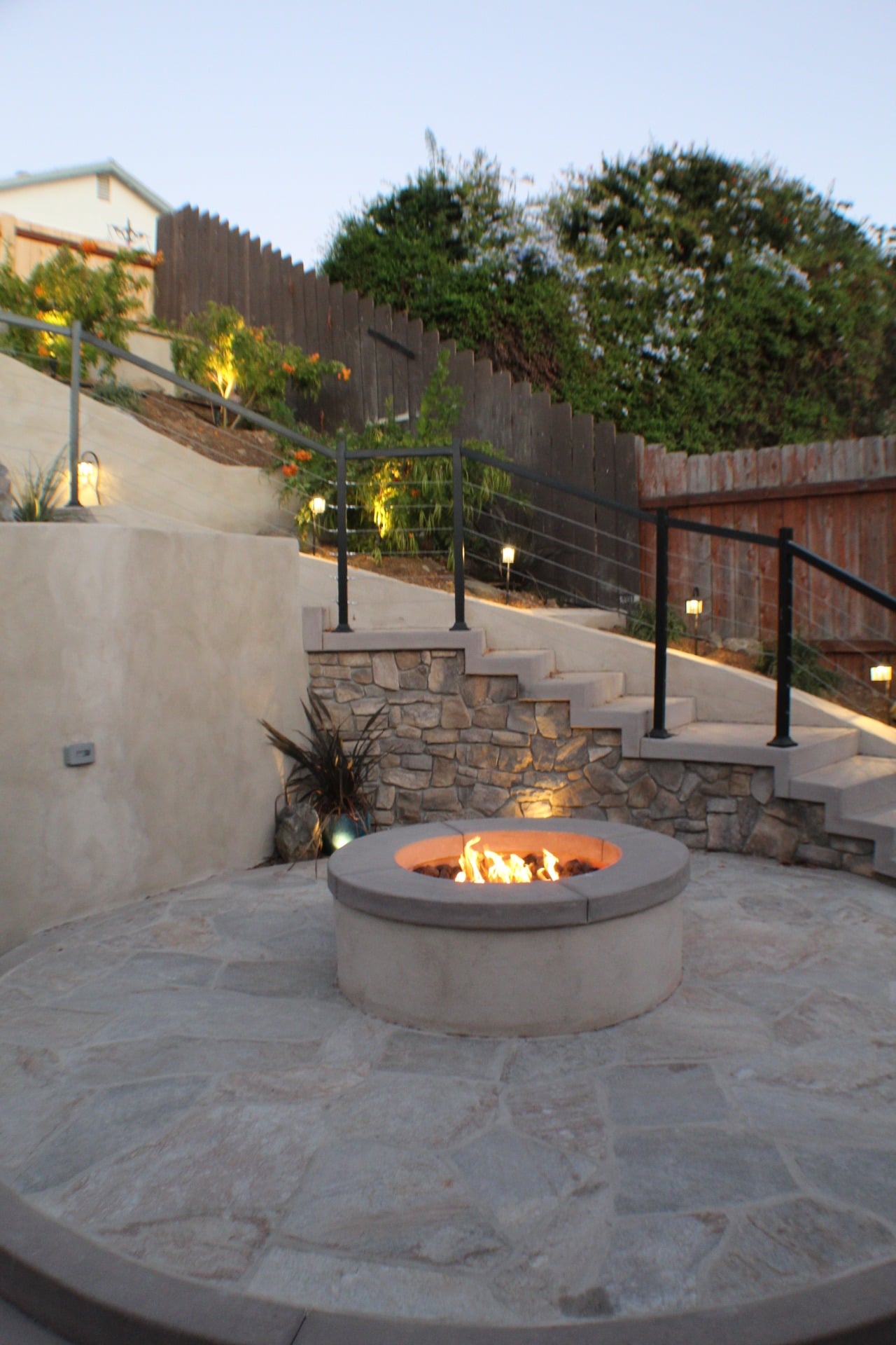 Lit fire pit on a flagstone patio with a stone-faced stair and cable rail rising behind it in Spring Valley