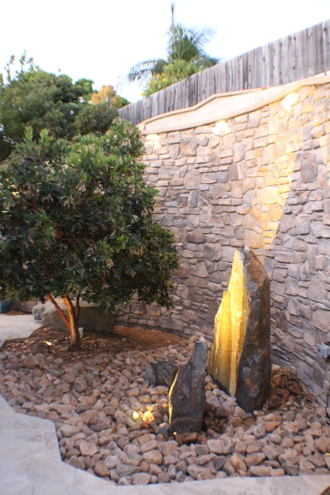 Uplit slate monoliths in a river-cobble bed against a stone-veneer retaining wall at dusk