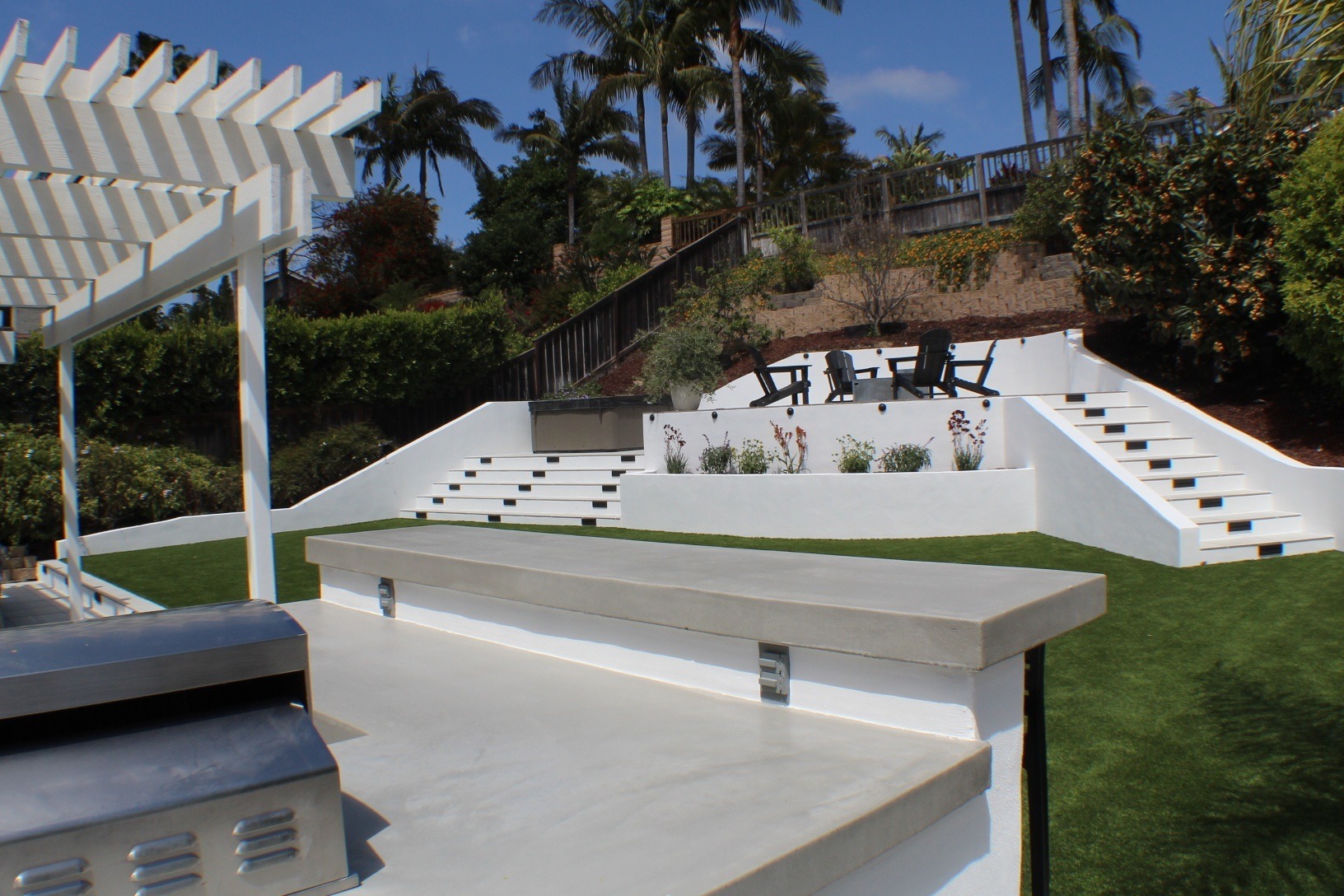 Poured-concrete outdoor kitchen counters under a white pergola beside terraced walls and turf