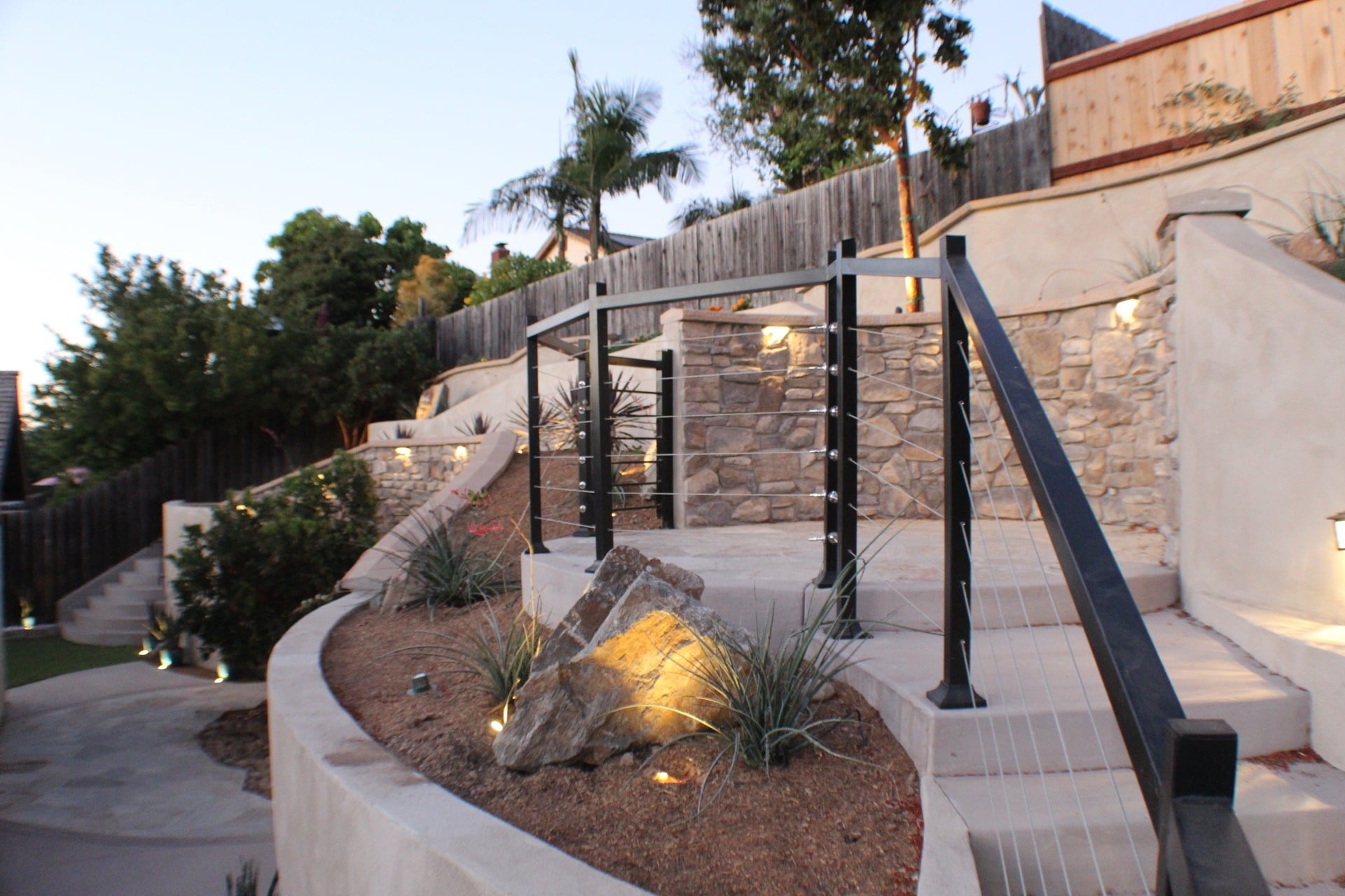 Stone-veneer retaining walls with cable railing and lit concrete steps at dusk